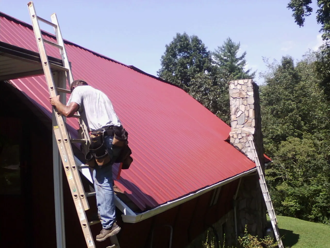 Roofer on a ladder beside a red metal roof and stone chimney — Soffit & Fascia Repair by Roofing Brothers in Mc Cracken